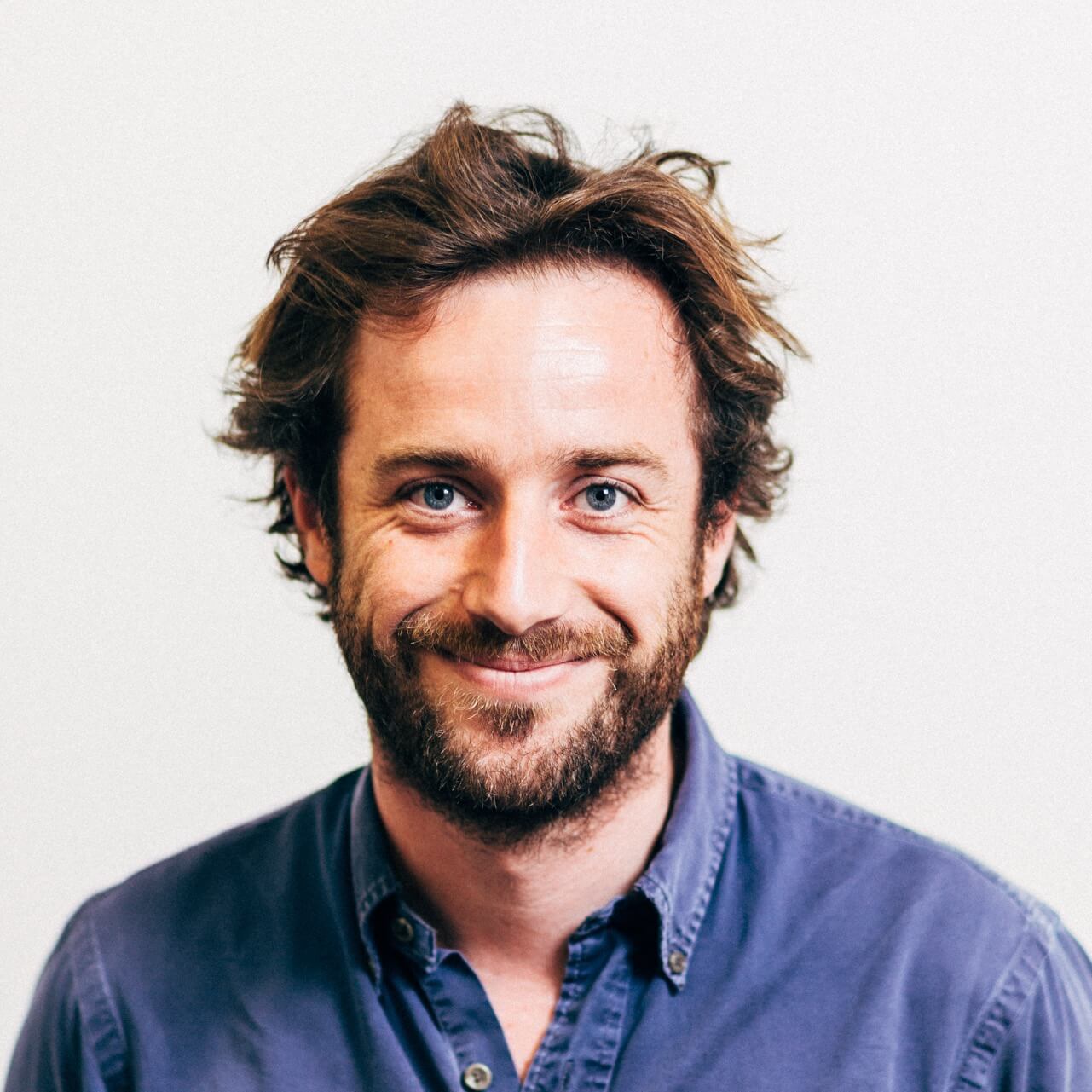 Smiling man with tousled brown hair and beard wearing a blue button-up shirt against a plain white background.