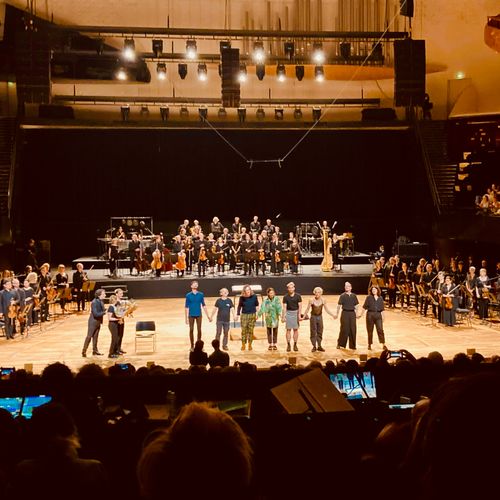 Orchestra and performers standing and holding hands on a stage during a curtain call in a concert hall.