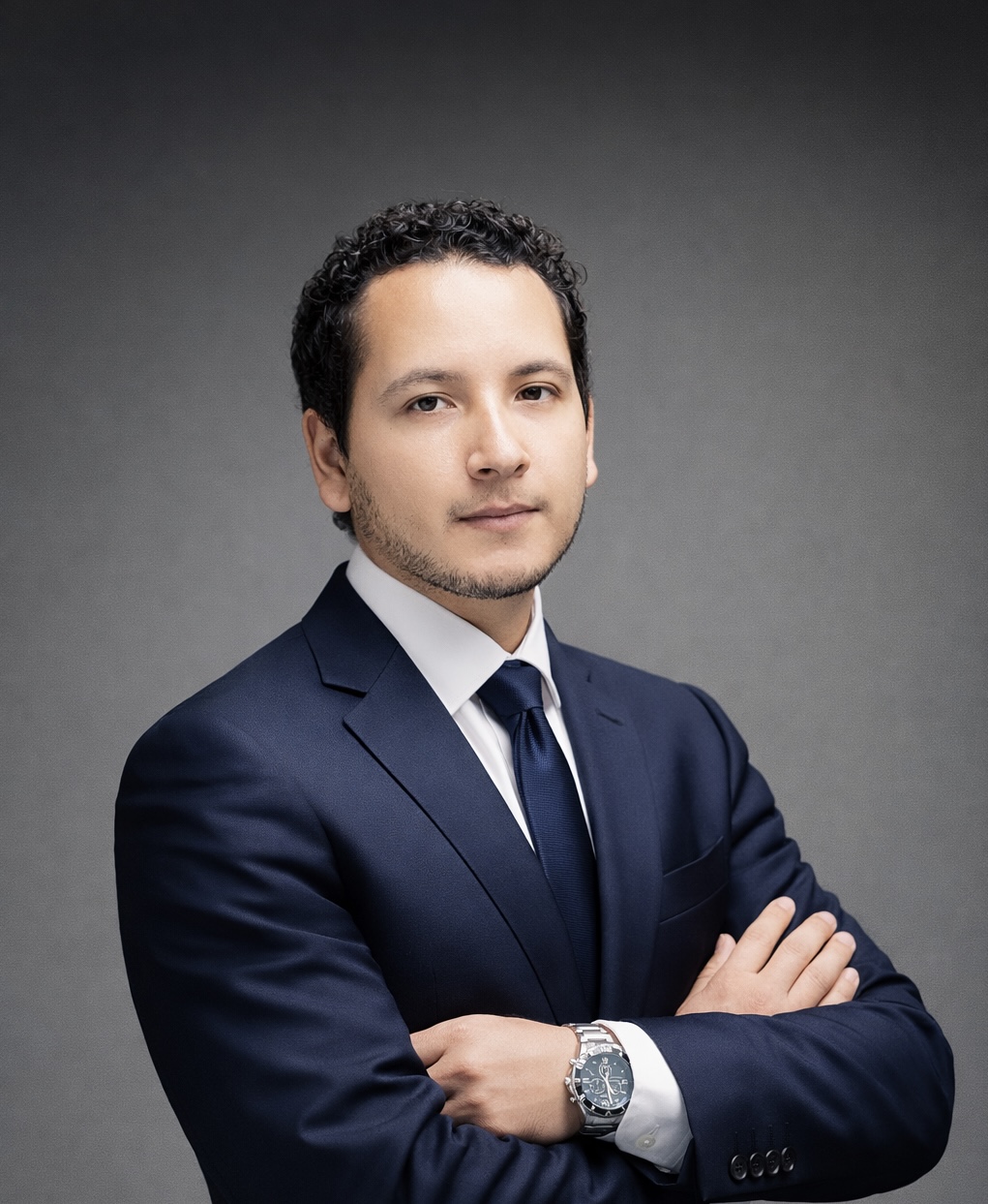 Confident man with curly hair wearing a navy suit and tie, arms crossed, against a gray background.