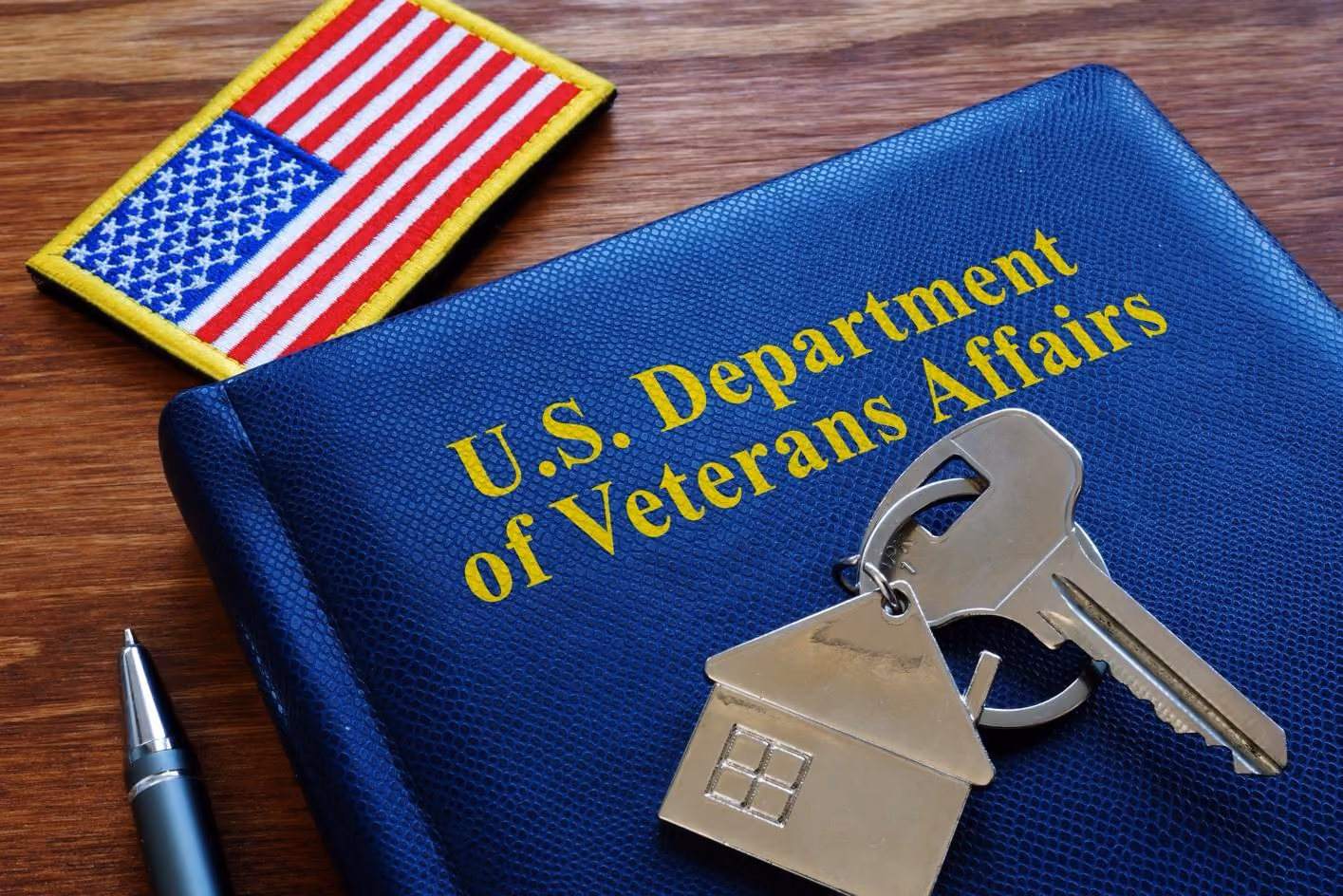 U.S. Department of Veterans Affairs notebook, U.S. flag patch, key with housing keychain, and pen on wood.