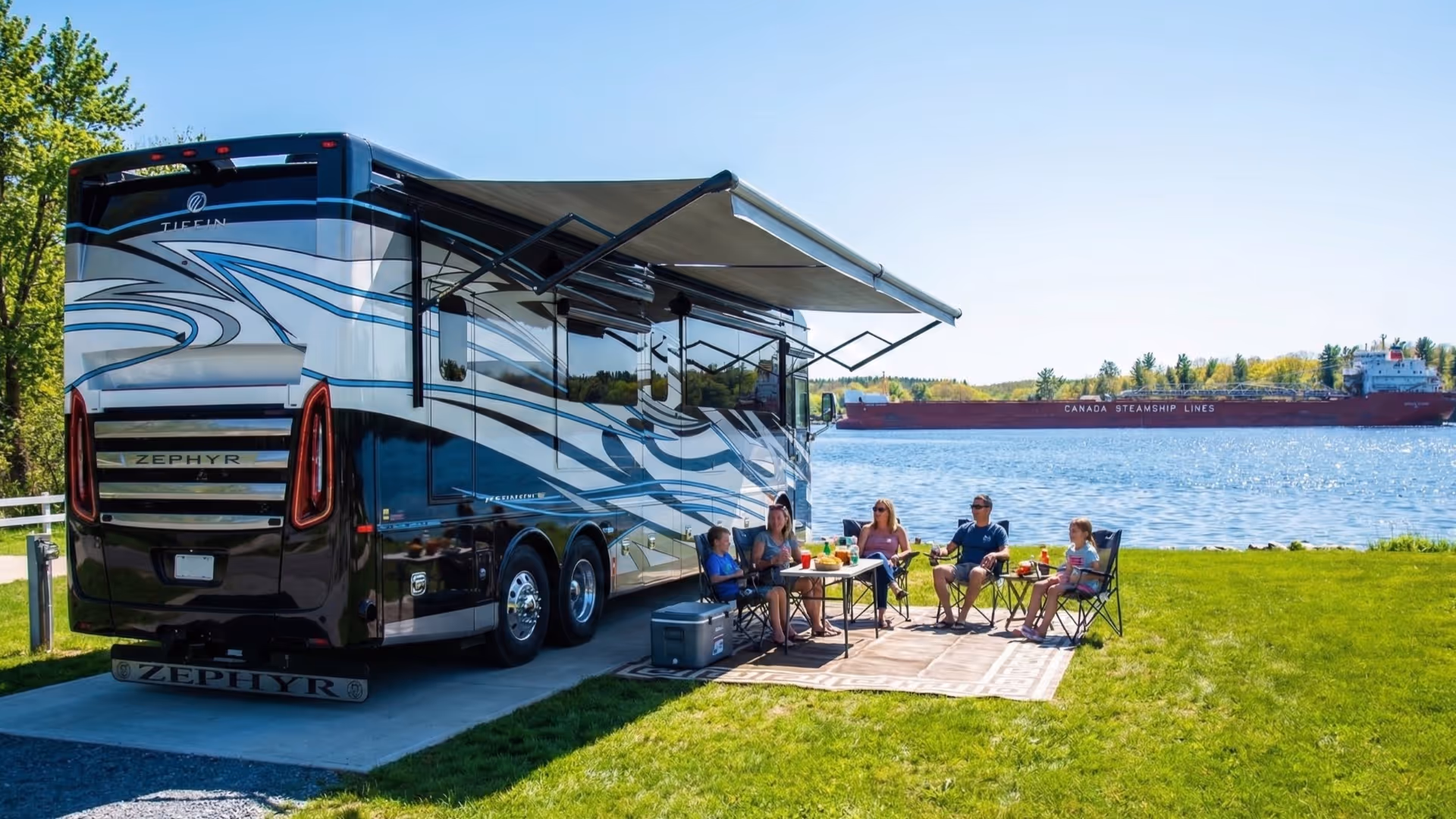 Family sitting outside RV under awning by water with a large ship in the background.