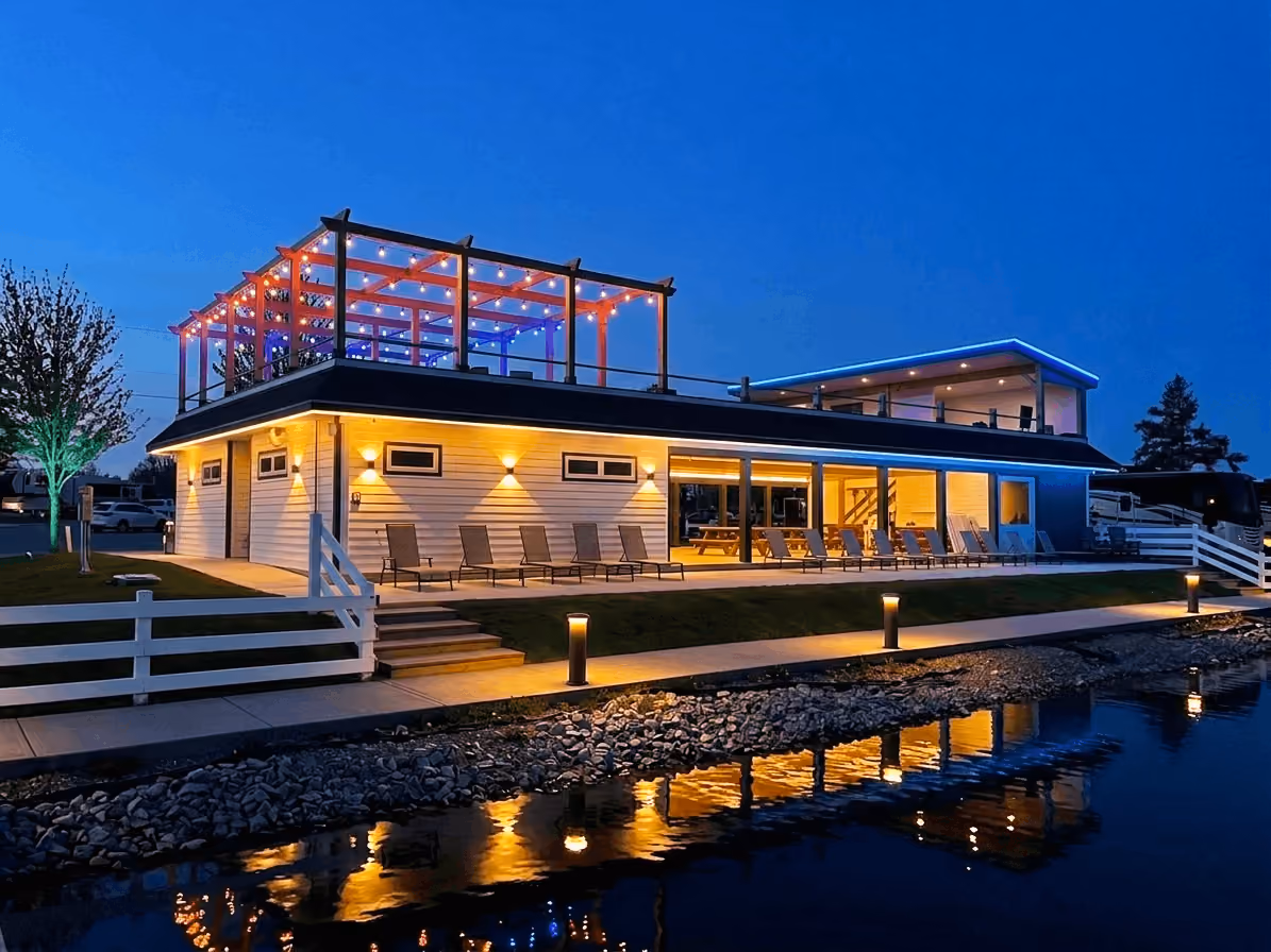Modern two-story building with string lights and lounge chairs, reflecting on water at dusk.