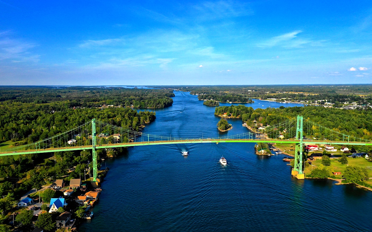 Aerial view of a green suspension bridge over a wide river with boats and forested riverbanks.