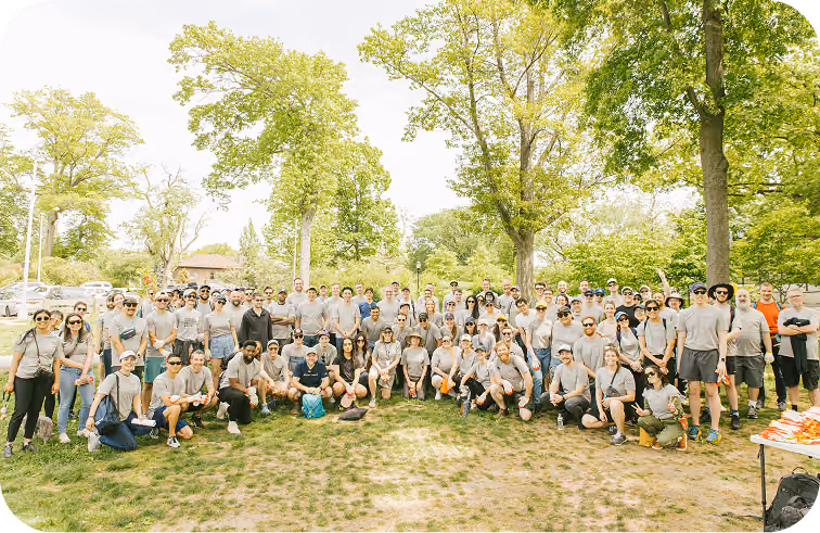 Large group of people wearing matching gray t-shirts gathered outdoors on grass with trees in the background.