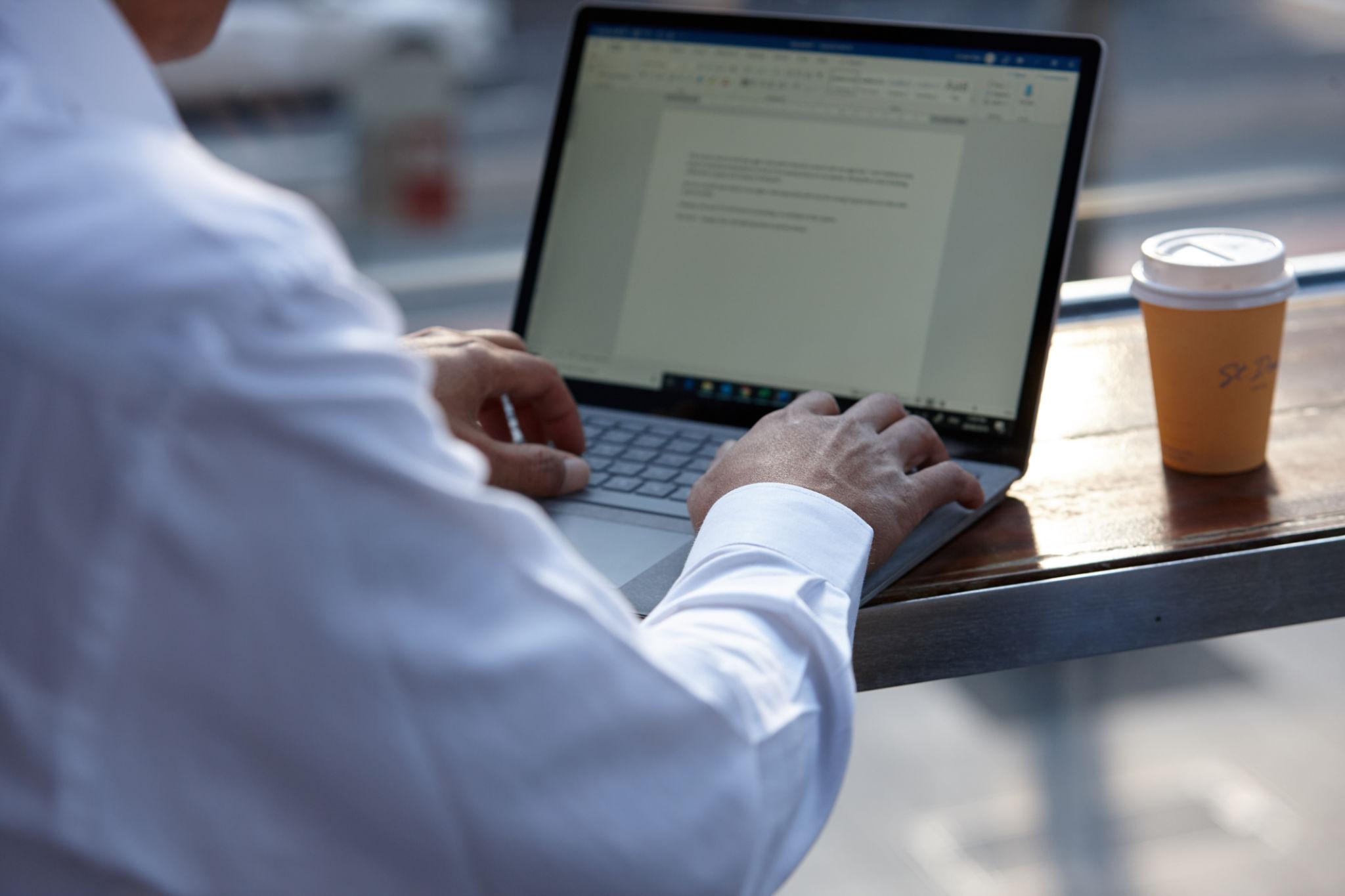 Close-up of hands typing on a laptop beside a takeaway coffee cup