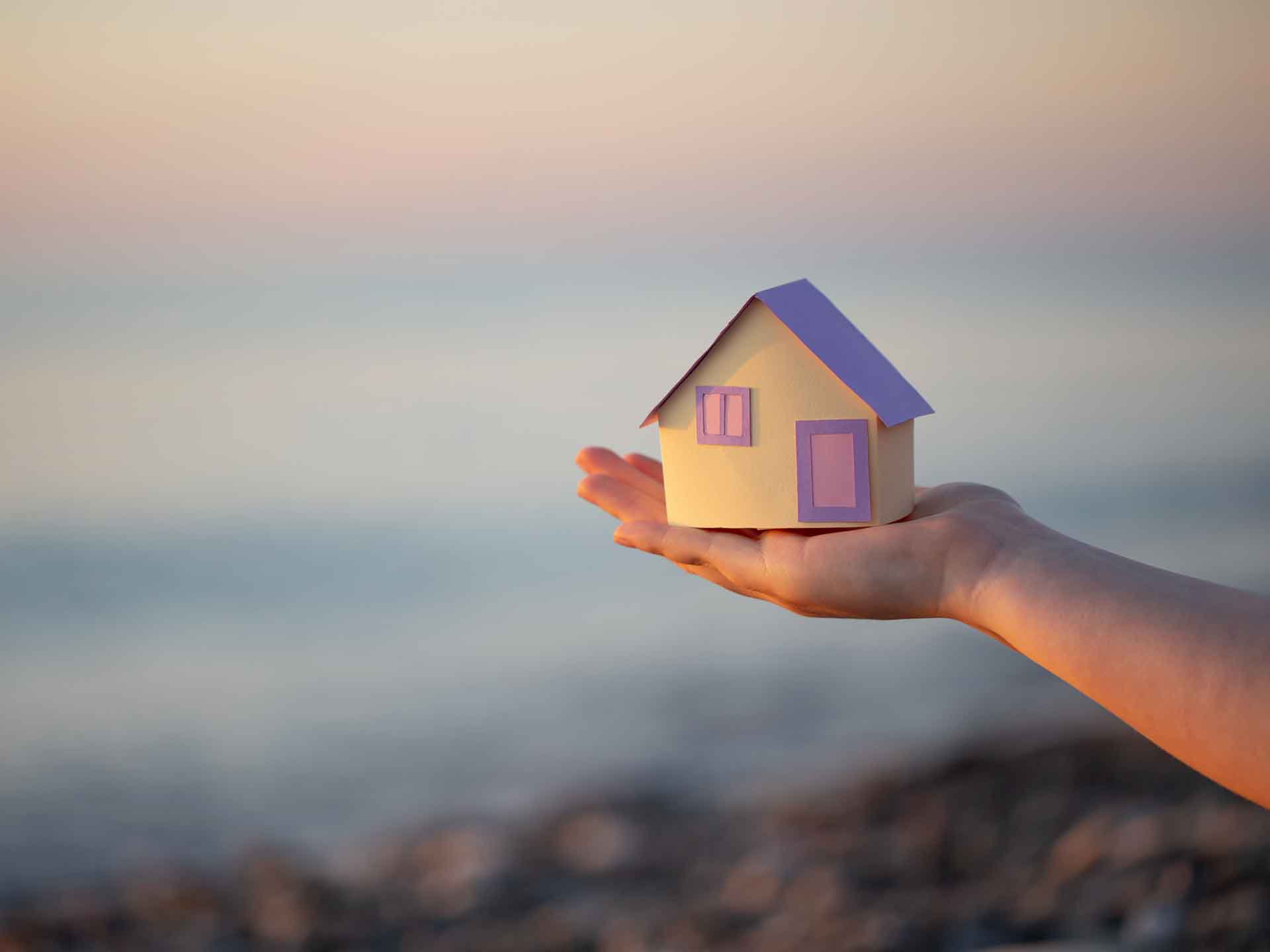 Hand holding a small paper house against a soft horizon