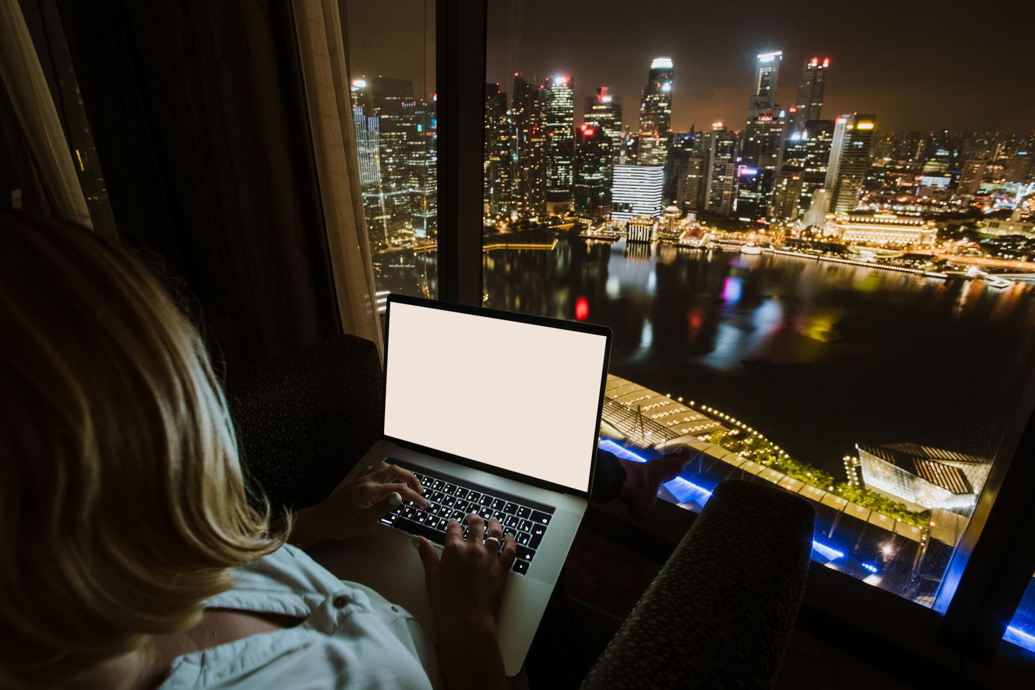 Woman using a laptop at night in front of a window overlooking a lit city skyline