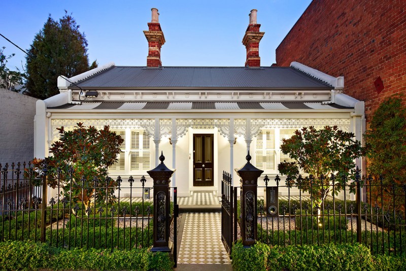 Heritage cottage with a lit veranda behind an iron fence at dusk