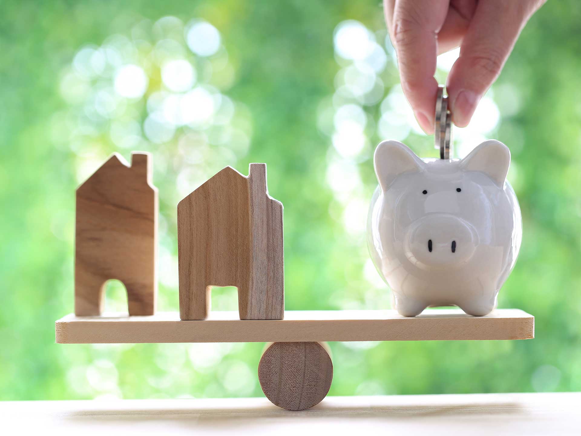 Wooden houses and a piggy bank balanced on a seesaw as a hand drops in a coin
