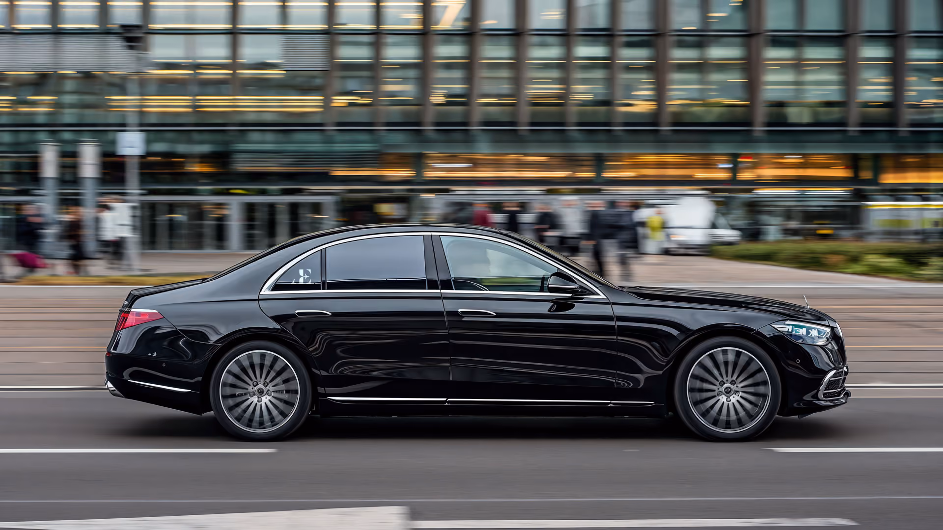 Een zwarte luxe sedan rijdt door een stadsstraat met op de achtergrond onscherpe voetgangers en een modern gebouw.