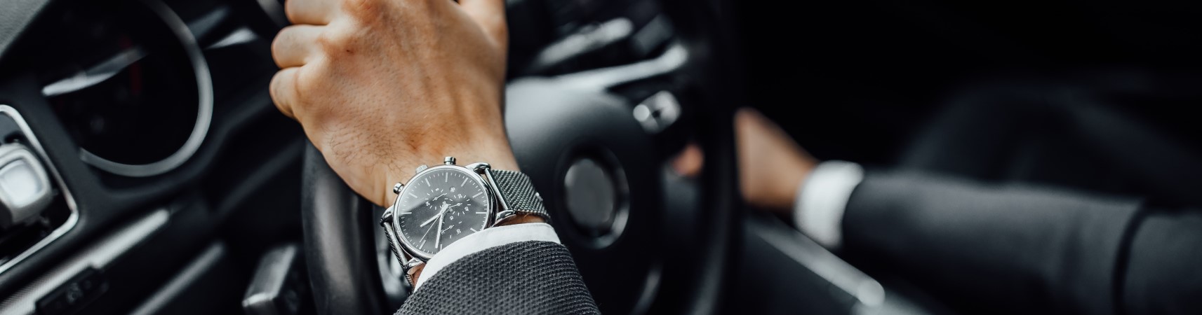 Close-up of a person wearing a wristwatch and suit driving a car, hands on the steering wheel.