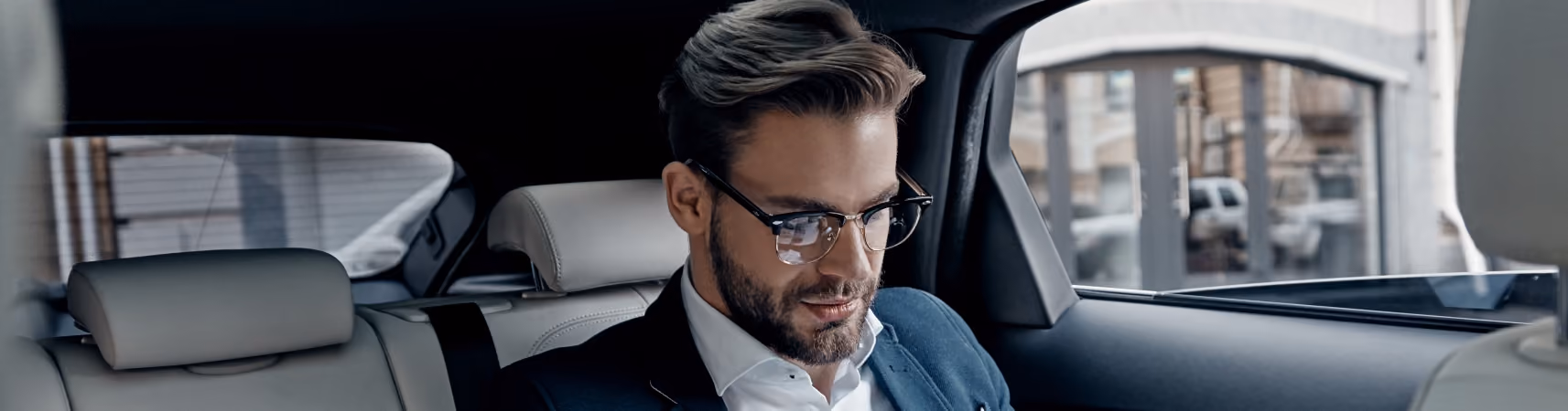 Man with glasses and beard dressed in a suit sitting in the back seat of a car looking down.