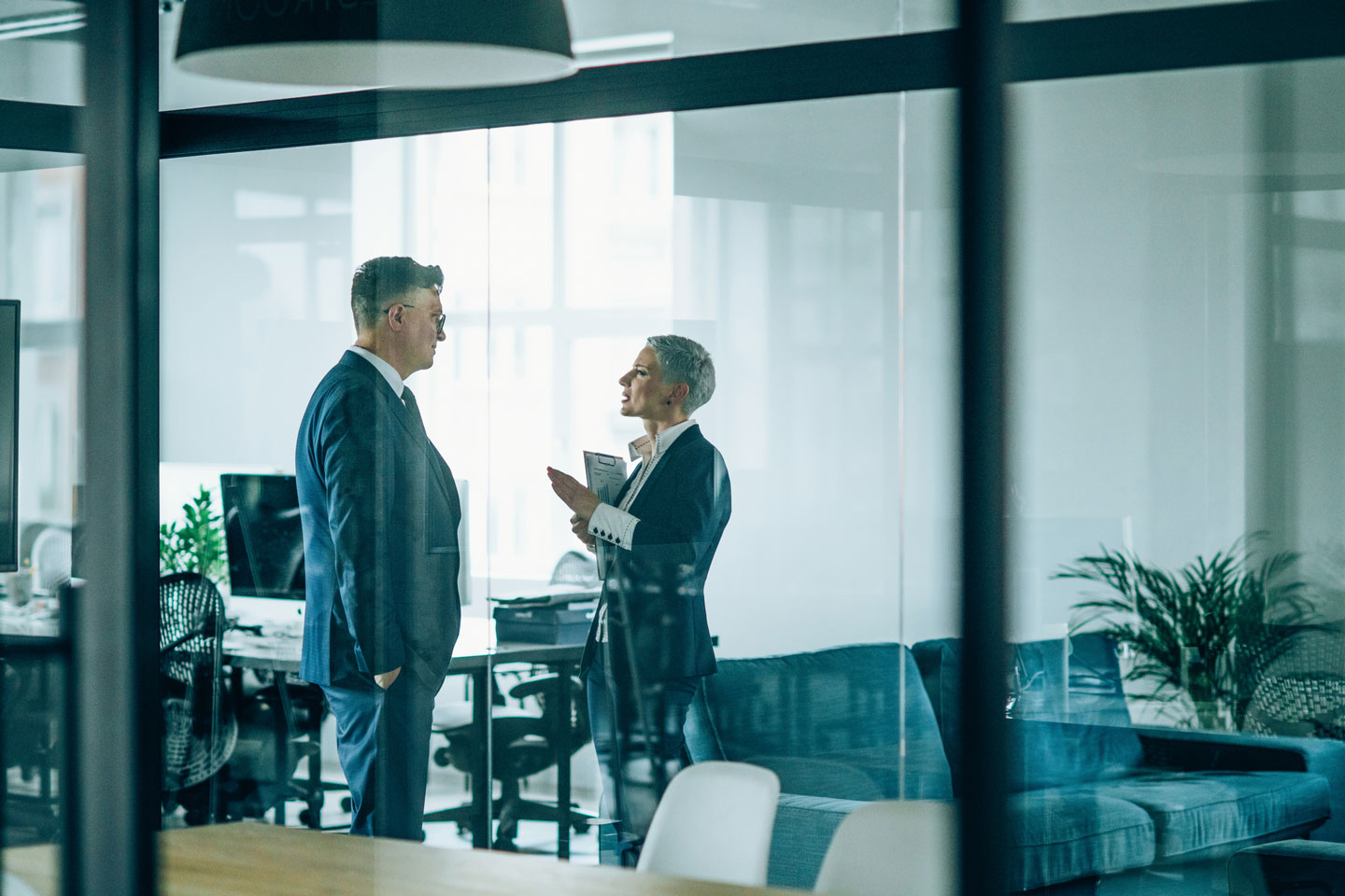 Two business professionals in formal attire discussing in a modern glass-walled office, representing corporate communication and professional collaboration.