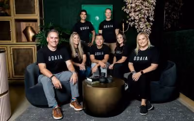 Group of seven people wearing black ODAIA shirts posing indoors around a round table with a green wall and decorative plants in the background.