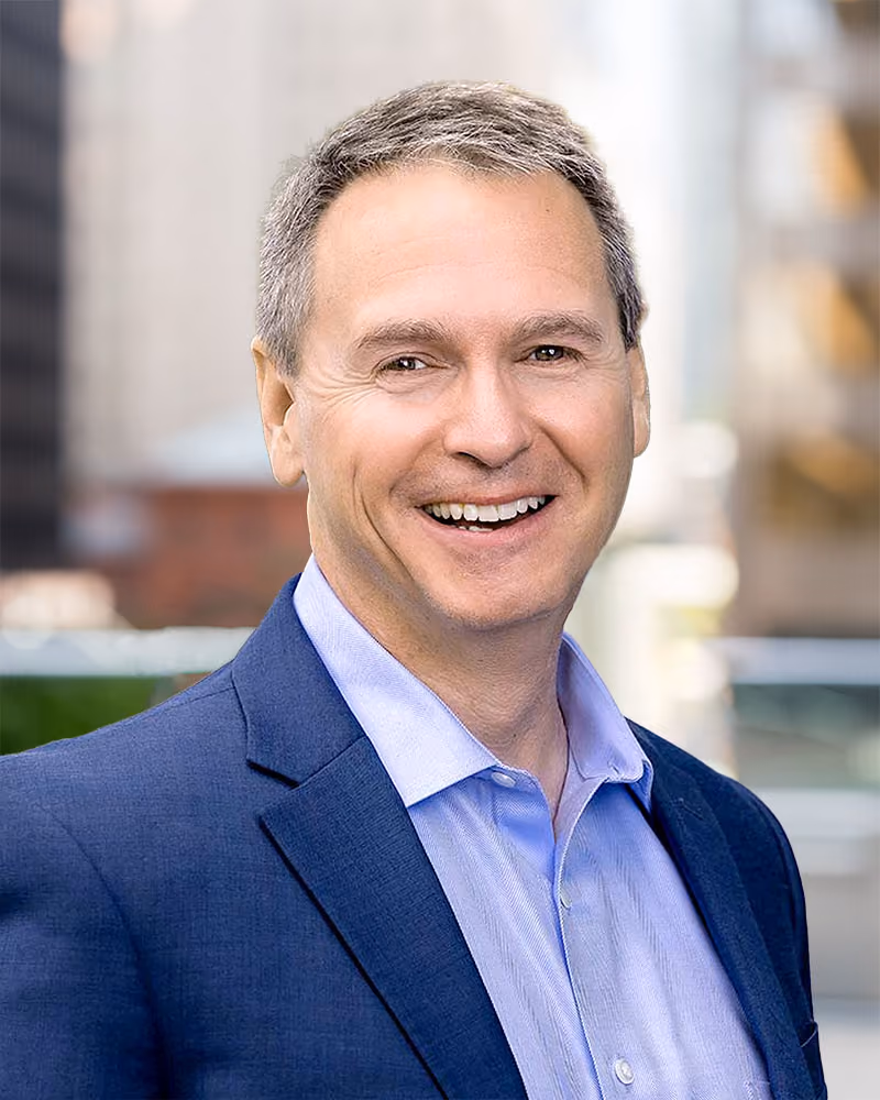 Smiling middle-aged man with short gray hair wearing a blue blazer and light blue shirt in an outdoor urban setting.