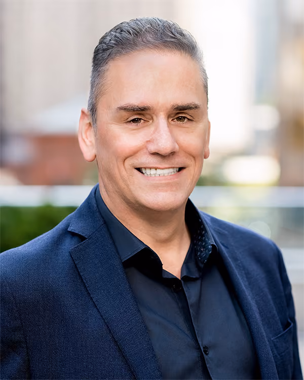 Smiling man wearing a navy blazer and black shirt with a blurred outdoor background.