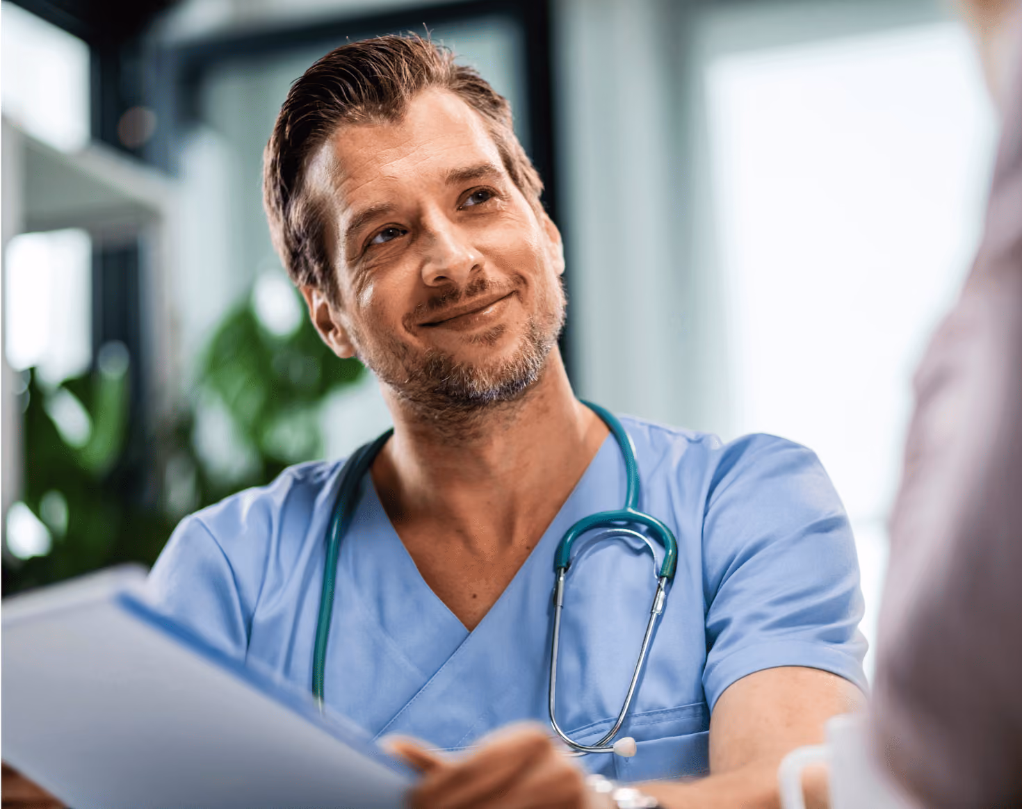 Male healthcare professional in blue scrubs with a stethoscope around his neck, smiling while holding a clipboard.
