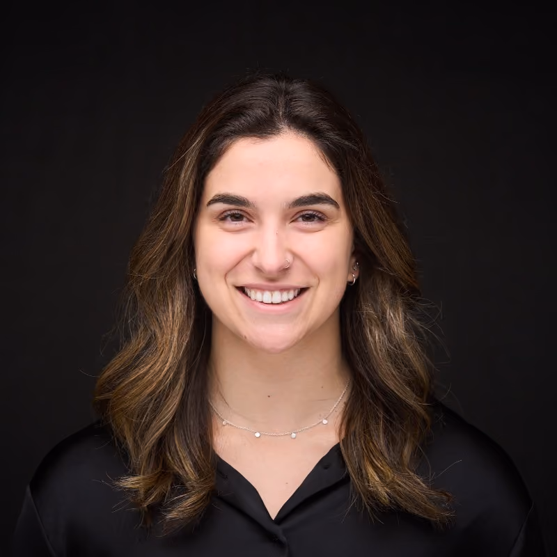 Smiling woman with shoulder-length brown hair wearing a black shirt and a delicate necklace against a black background.