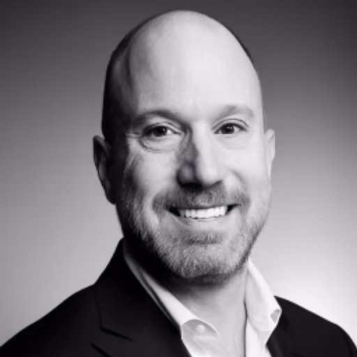 Smiling bald man with a beard wearing a dark blazer and white shirt against a gradient background.