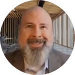 Smiling bald man with a long gray beard wearing a suit jacket in a wooden building with large windows.