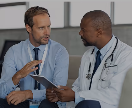 A doctor in white coat with a stethoscope consulting attentively with a man in a blue shirt holding a tablet.