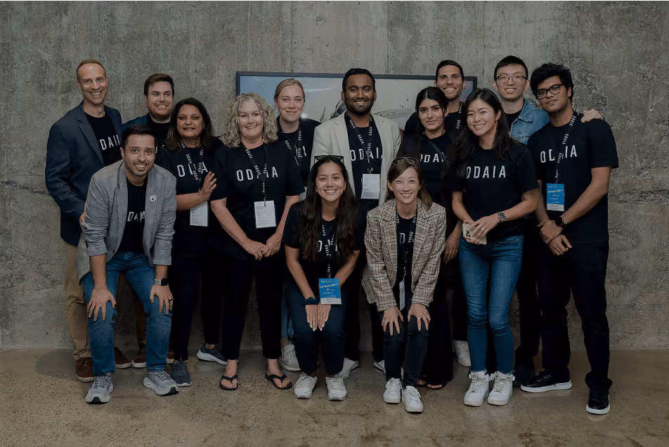 Group of 14 smiling people posing indoors against a concrete wall, many wearing black ODAIA t-shirts and some with conference badges.
