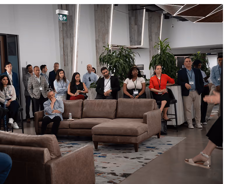 Group of diverse people attentively listening in a modern indoor space near a brown sectional sofa and large plants.