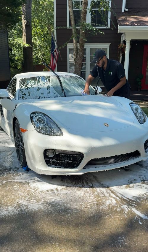 Man washing a white Porsche sports car covered in soap suds on a driveway in front of a house.