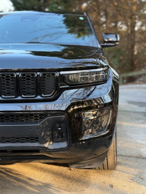Close-up of the front right side of a black SUV with detailed headlight and grille, parked on a paved road with autumn trees in the background.