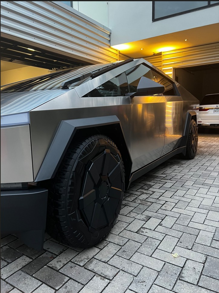Silver Tesla Cybertruck parked on a gray brick driveway in front of a modern house garage.