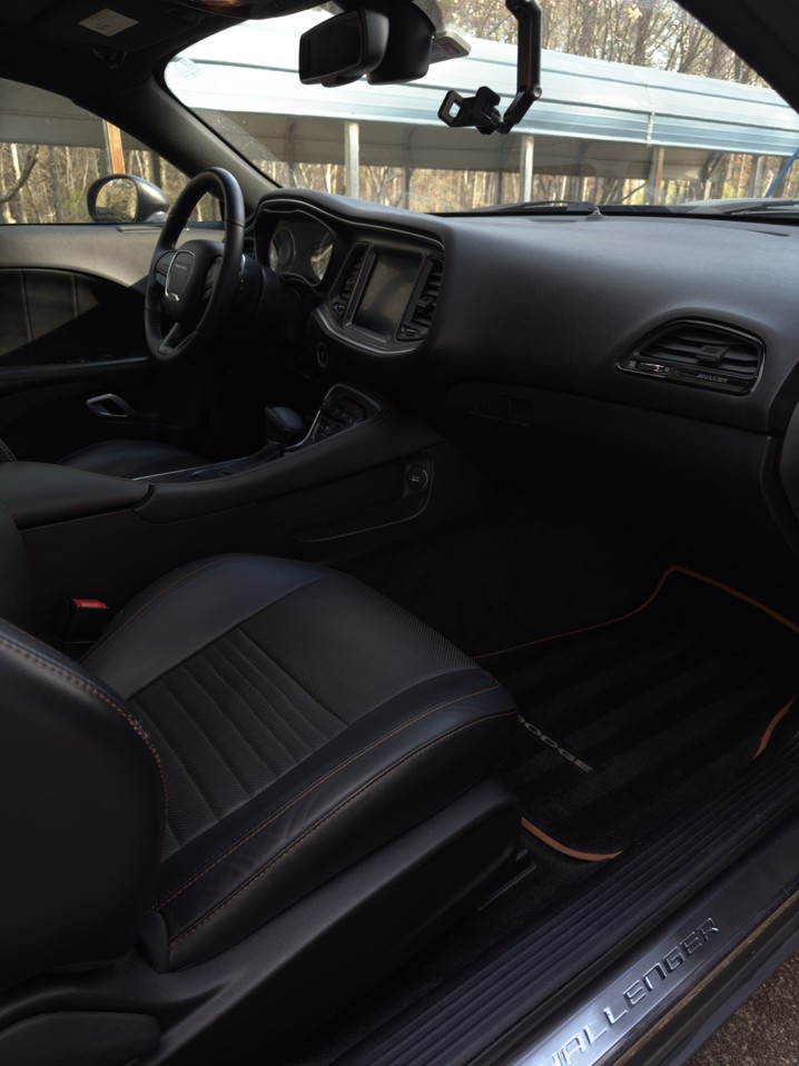 Interior of a Dodge Challenger showing the black leather driver's seat, steering wheel, dashboard, and console with woodsy background visible through windows.