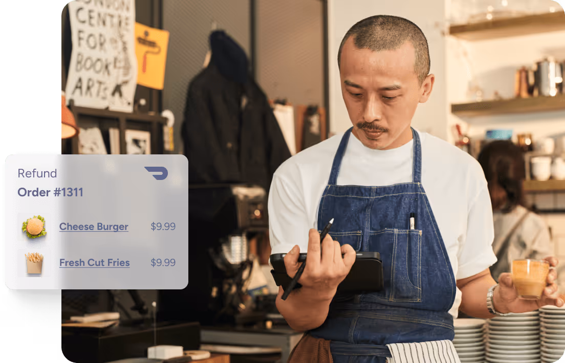 Barista in a denim apron holding a small cup of coffee and checking a tablet with a refund order showing cheese burger and fresh cut fries priced at $9.99 each.