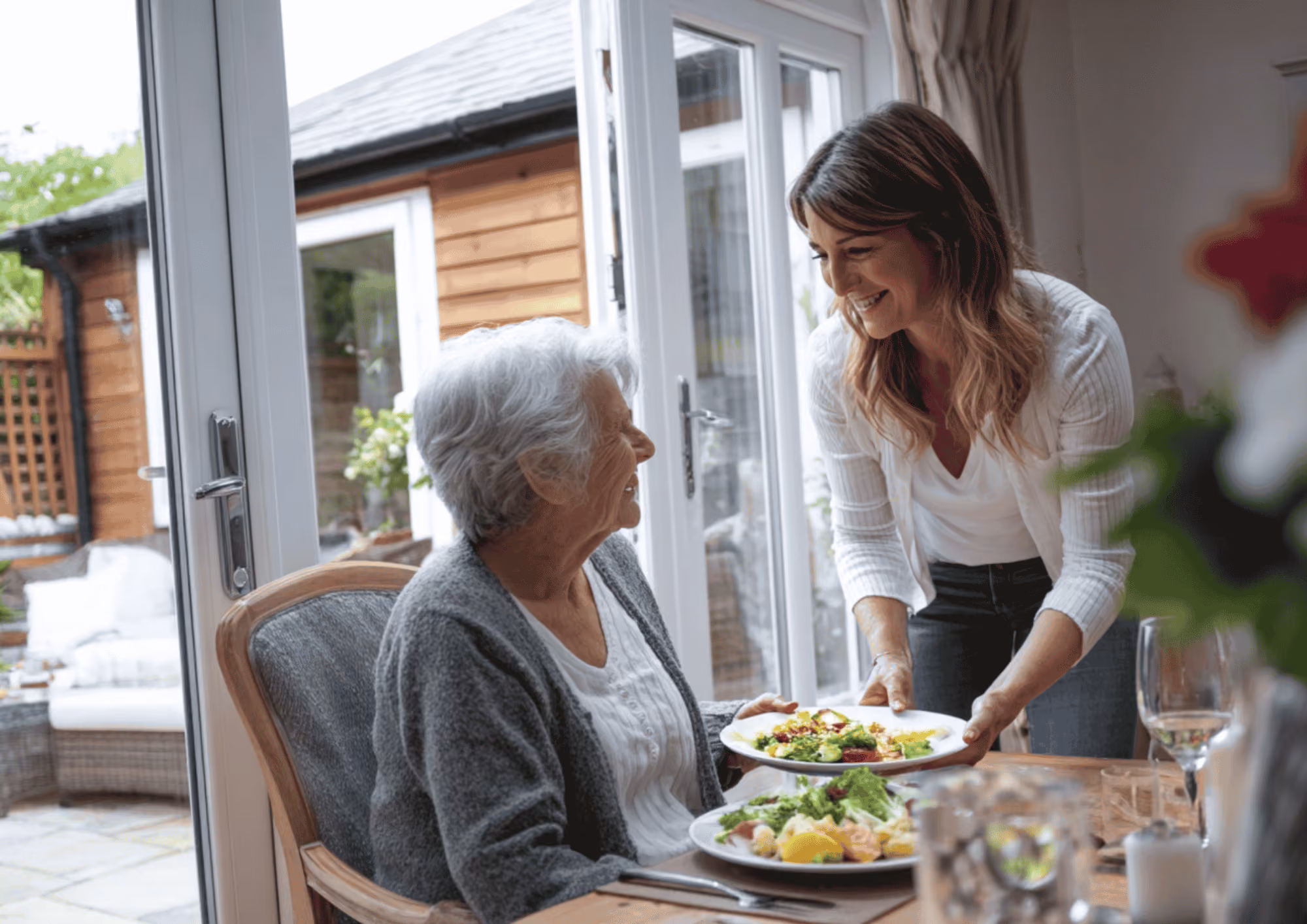 A young woman serving a plate of food to an elderly woman seated at a dining table near large glass doors, both smiling warmly at each other.