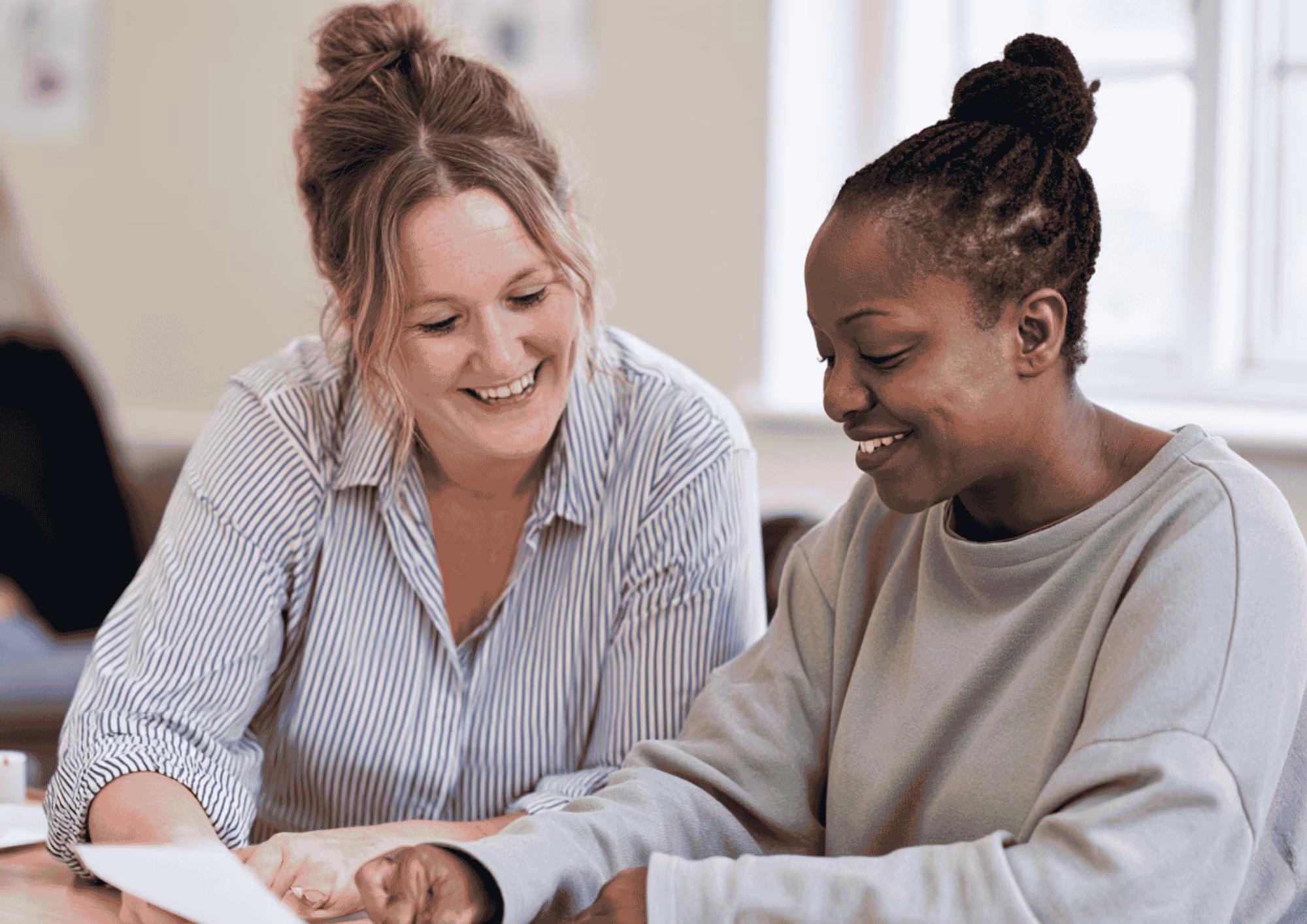 Two women sitting together at a table, smiling and looking at papers in front of them as they collaborate.
