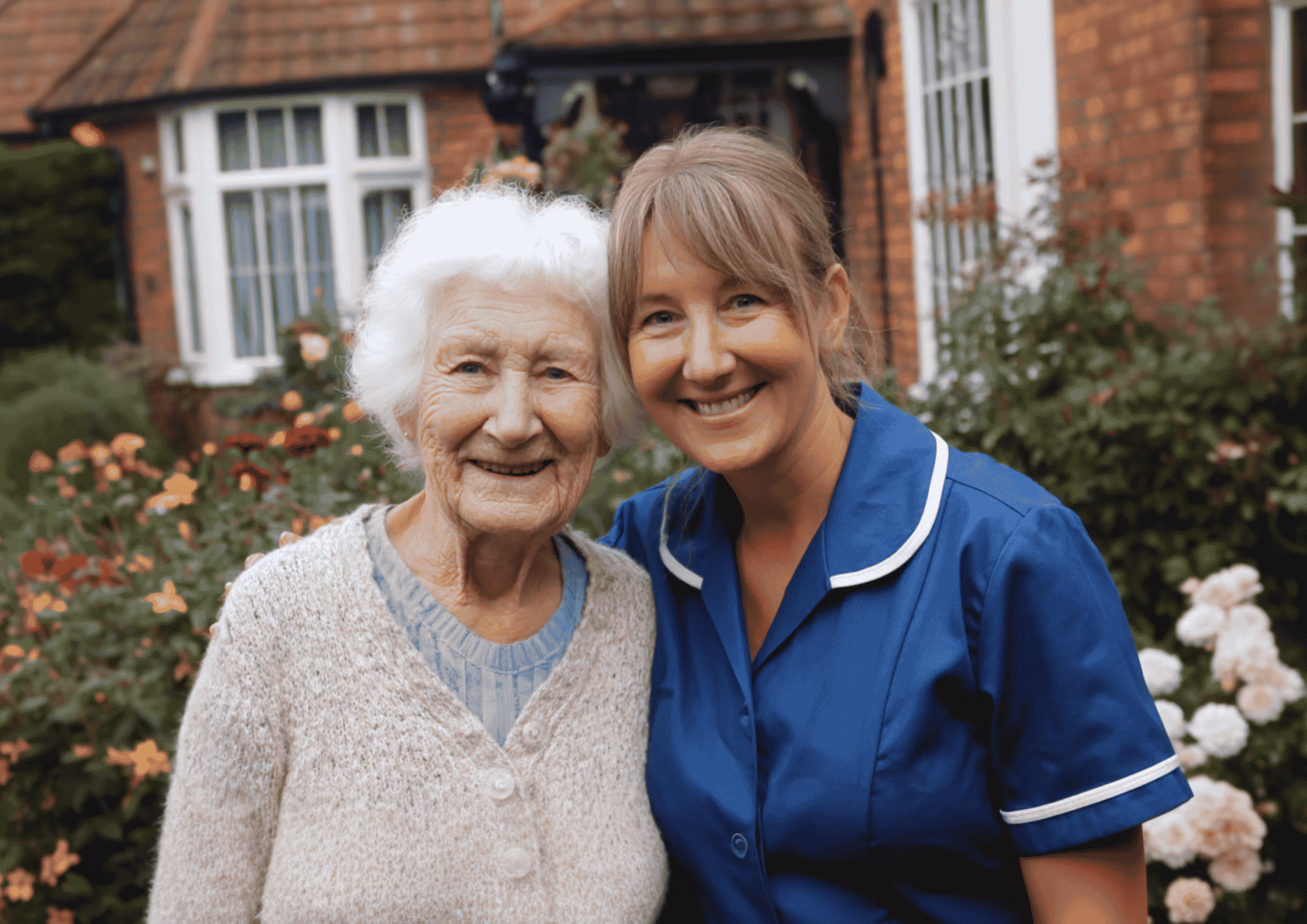 An elderly woman with white hair in a beige cardigan stands smiling next to a smiling female caregiver wearing a blue uniform outside a brick house with flowers and greenery.