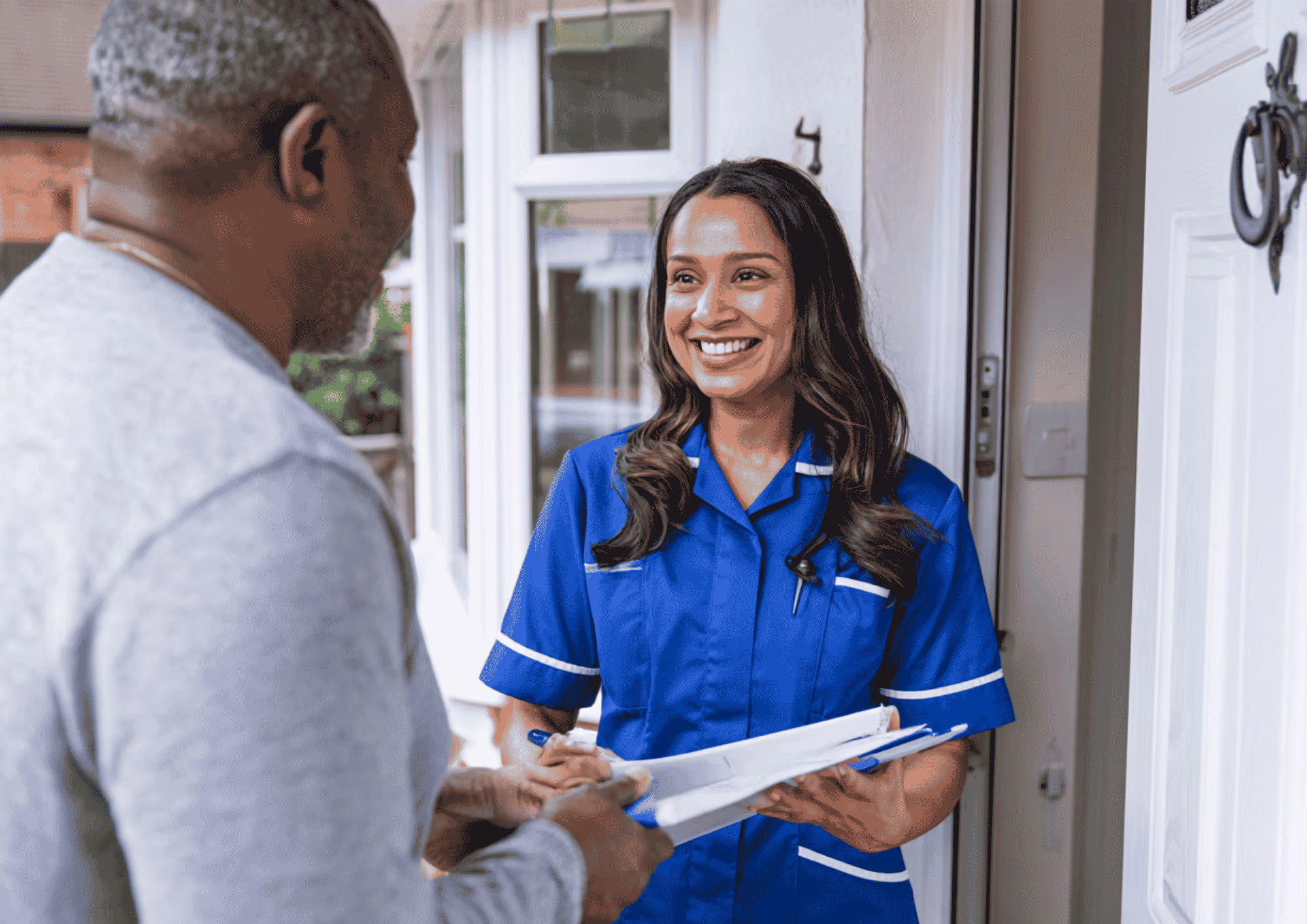 Smiling female nurse in blue uniform holding a clipboard talks to an older man at the door of a house.