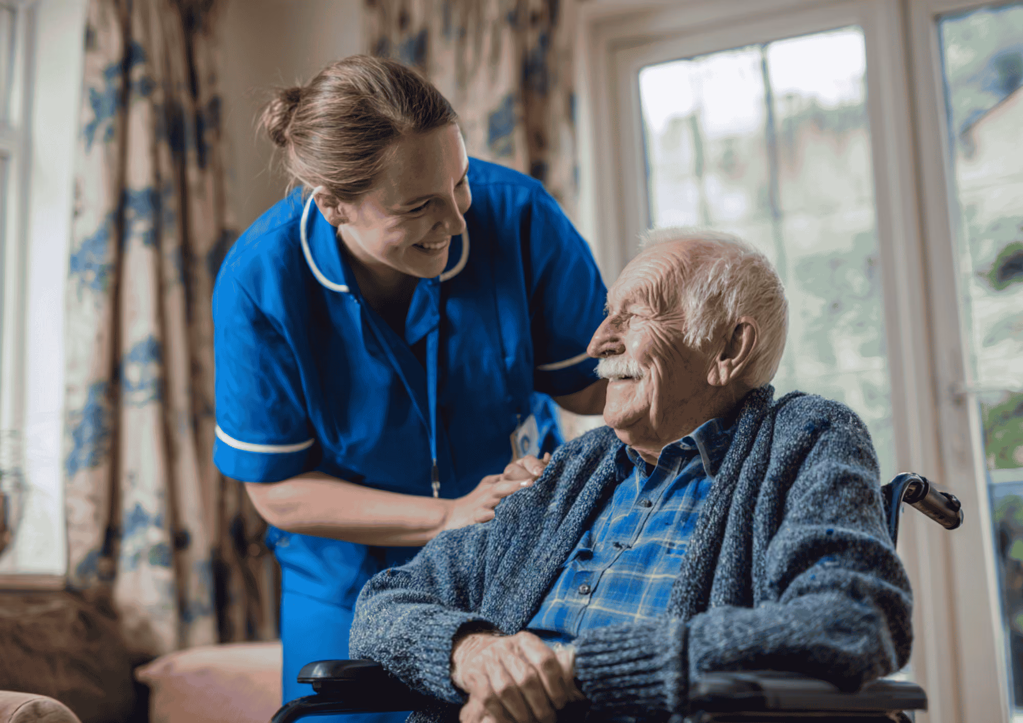 A smiling nurse in blue scrubs leaning over and engaging kindly with an elderly man in a wheelchair wearing a blue plaid shirt and gray cardigan, inside a bright room with large windows and patterned curtains.
