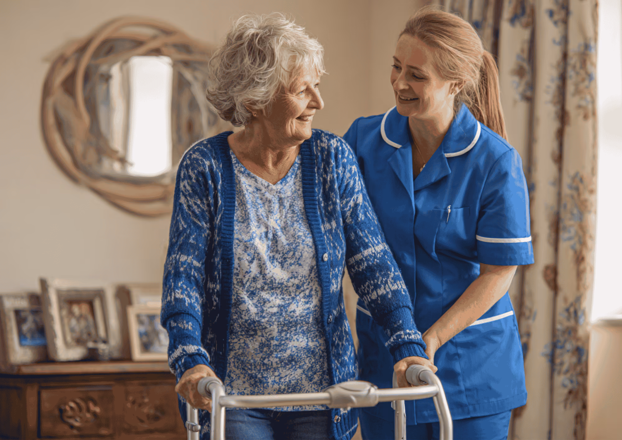A smiling elderly woman using a walker is being assisted by a female caregiver in blue uniform in a cozy, well-lit room with framed photos and decorative curtains.