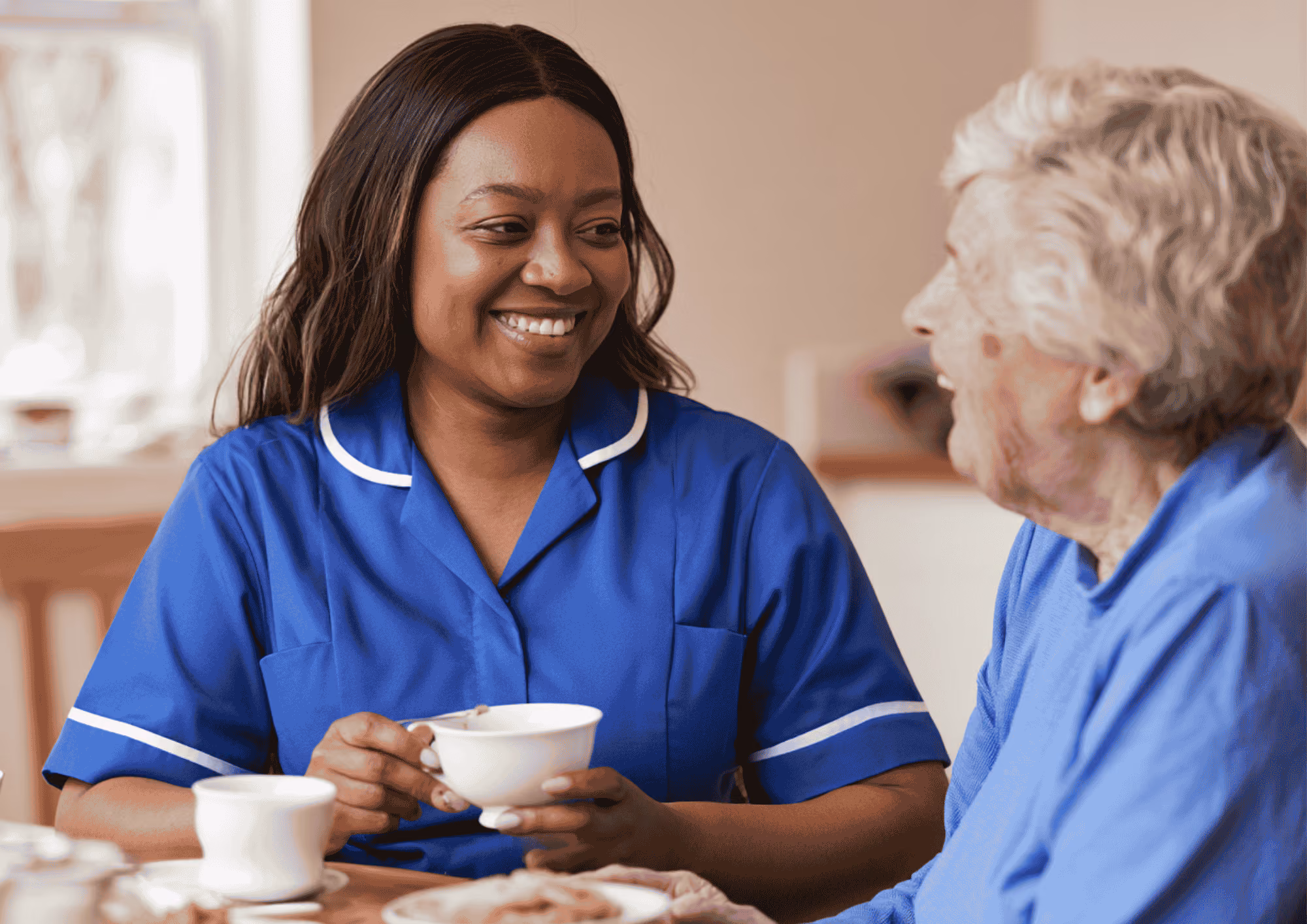 Smiling female caregiver in blue uniform holding a teacup, sitting at a table and talking with an elderly woman in a blue sweater.