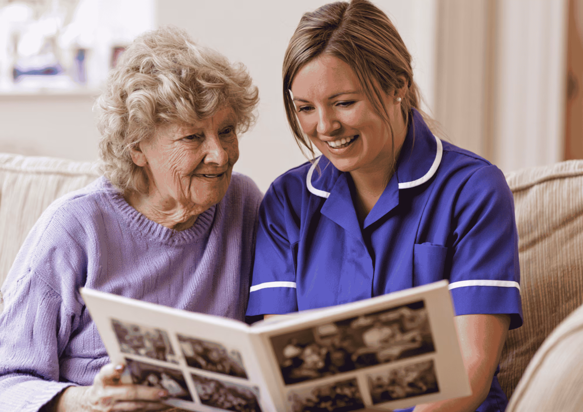 An elderly woman and a caregiver in a purple uniform sitting on a couch together, smiling and looking at a photo album.