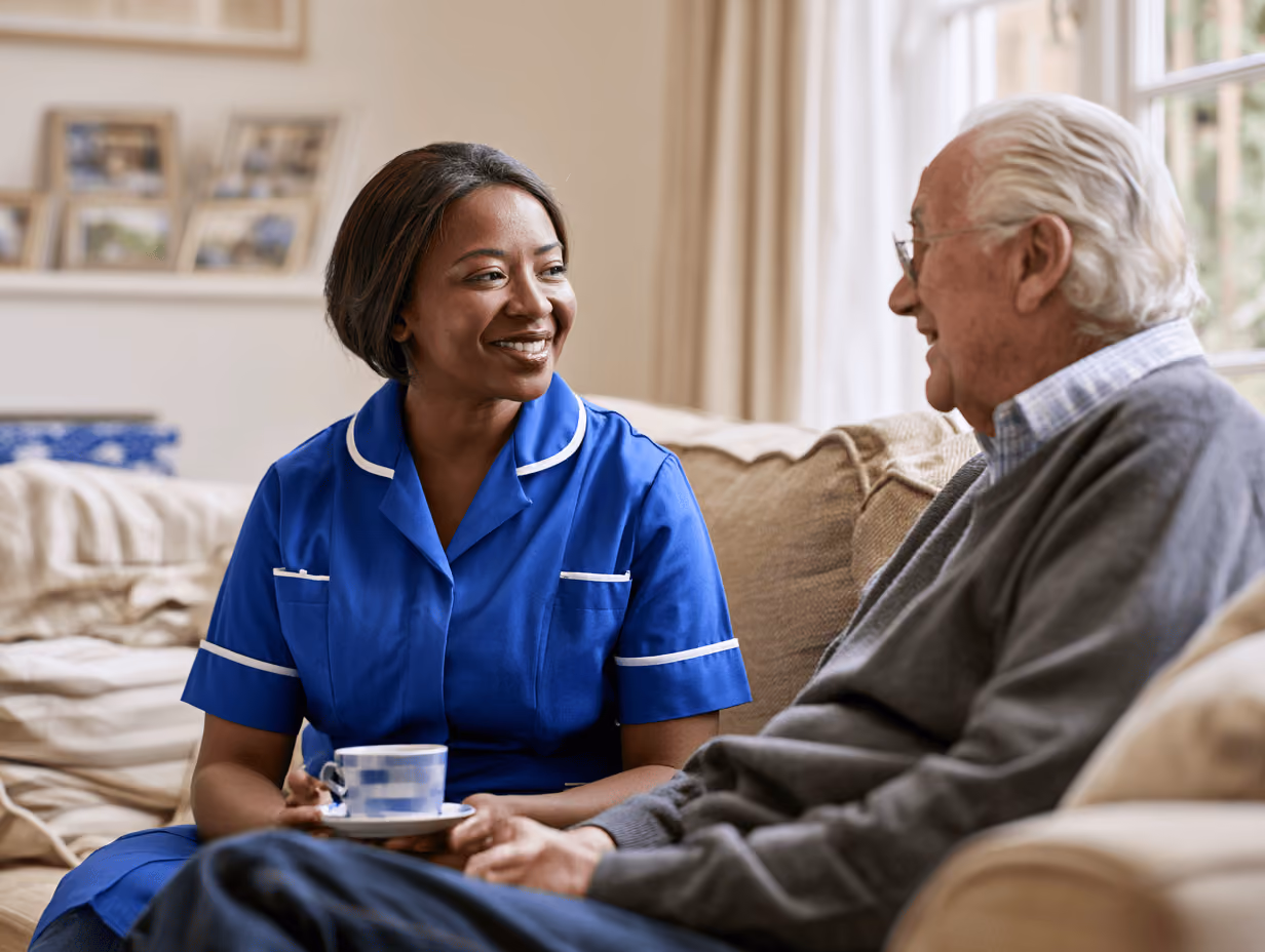 A smiling female caregiver in blue uniform sitting on a couch, holding a cup and saucer, talking with an elderly man wearing glasses and a gray sweater.