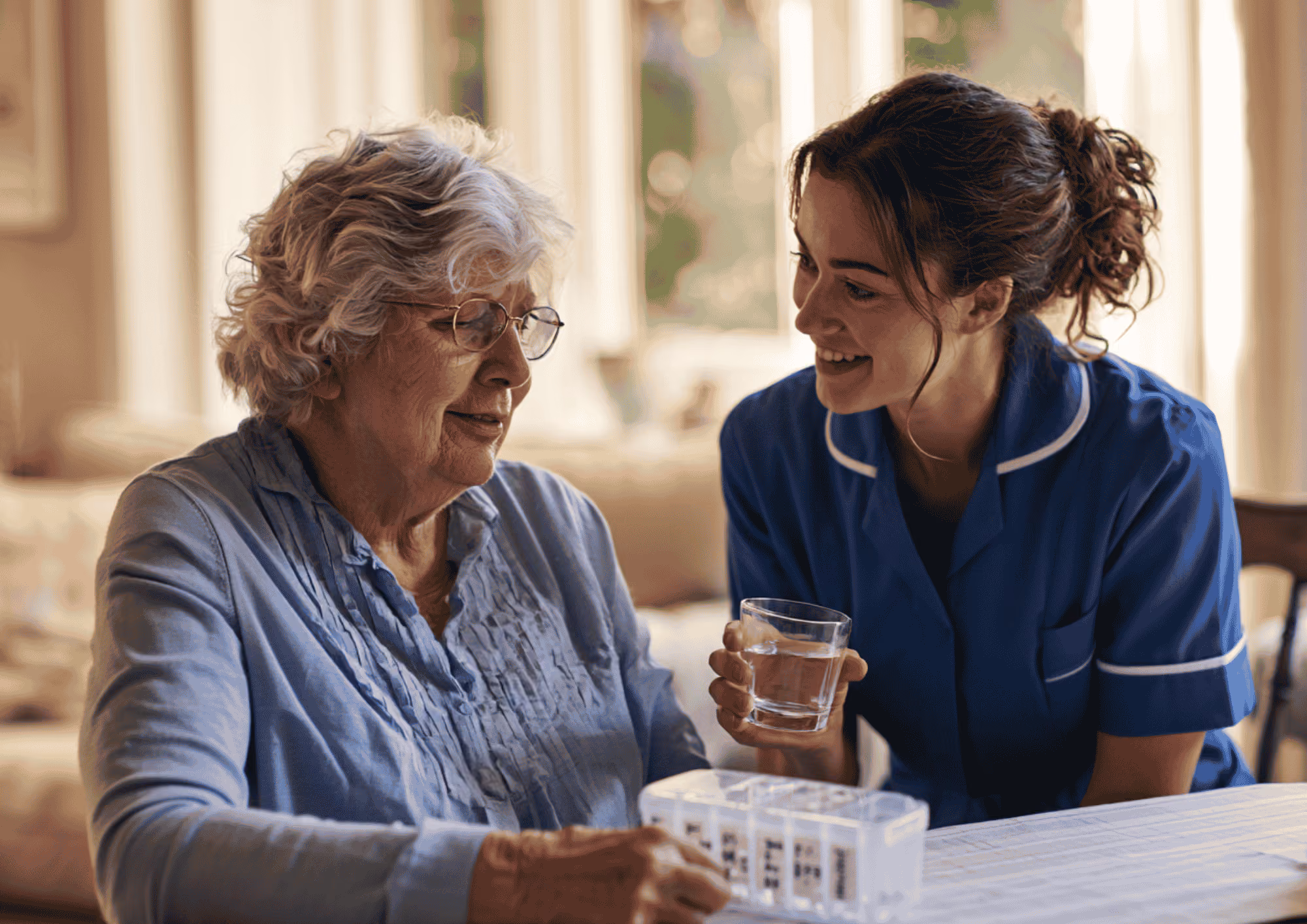 Elderly woman sitting at a table with a weekly pill organizer while a smiling caregiver in a blue uniform offers her a glass of water in a warmly lit room.