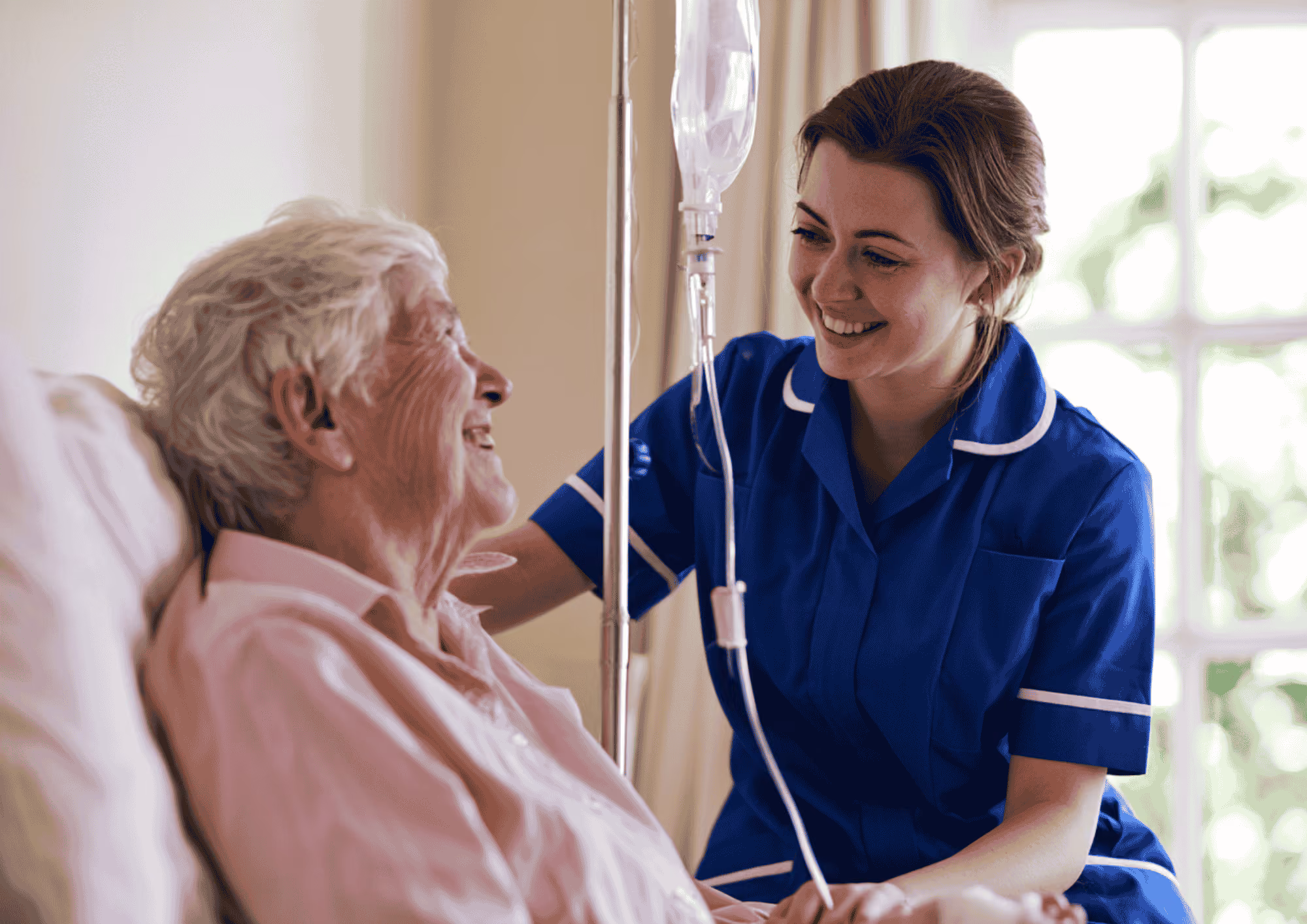 A smiling care worker in a blue uniform supporting an elderly man receiving intravenous treatment at home.