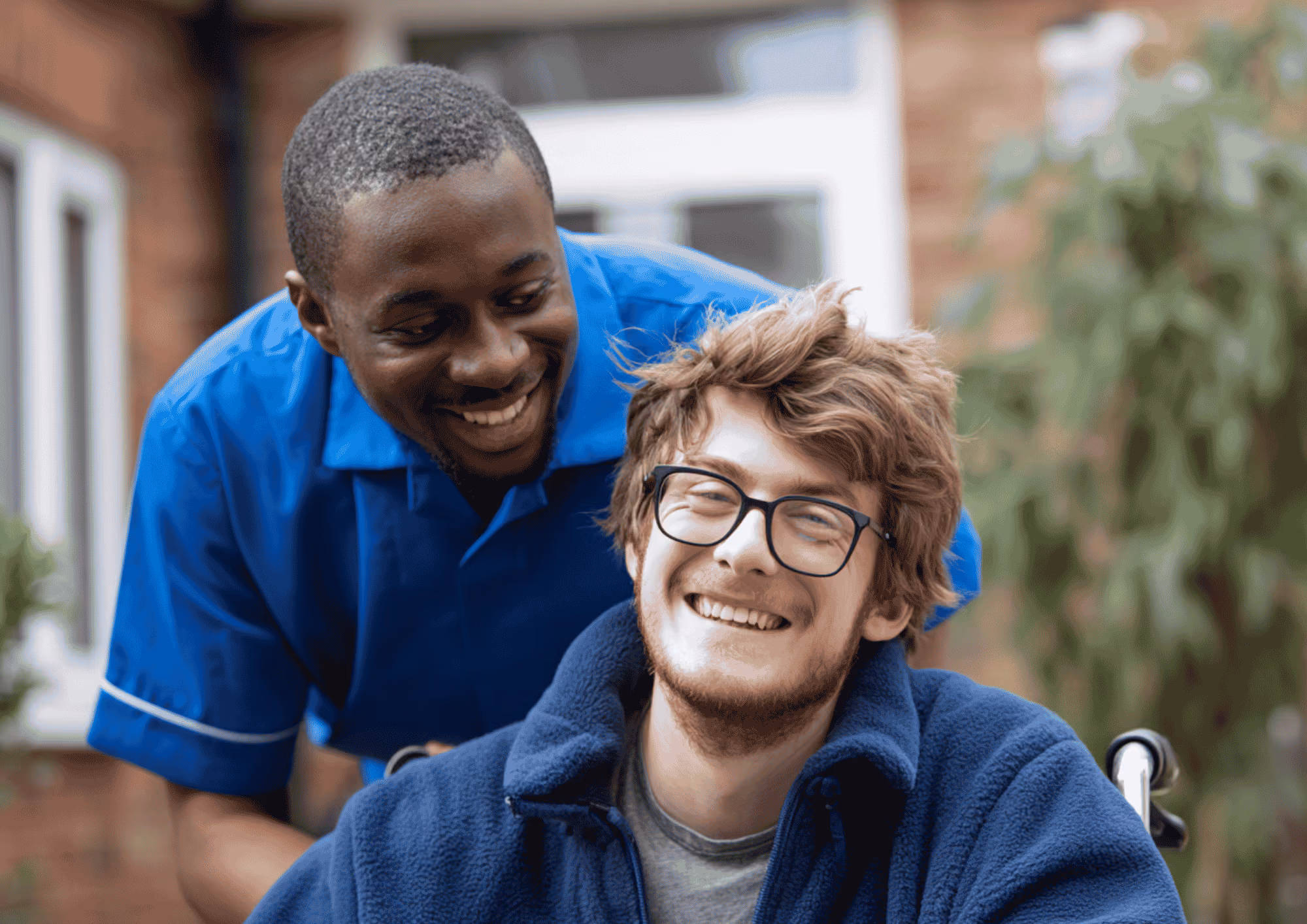 A smiling care worker in a blue uniform supporting a young man using a wheelchair outdoors.