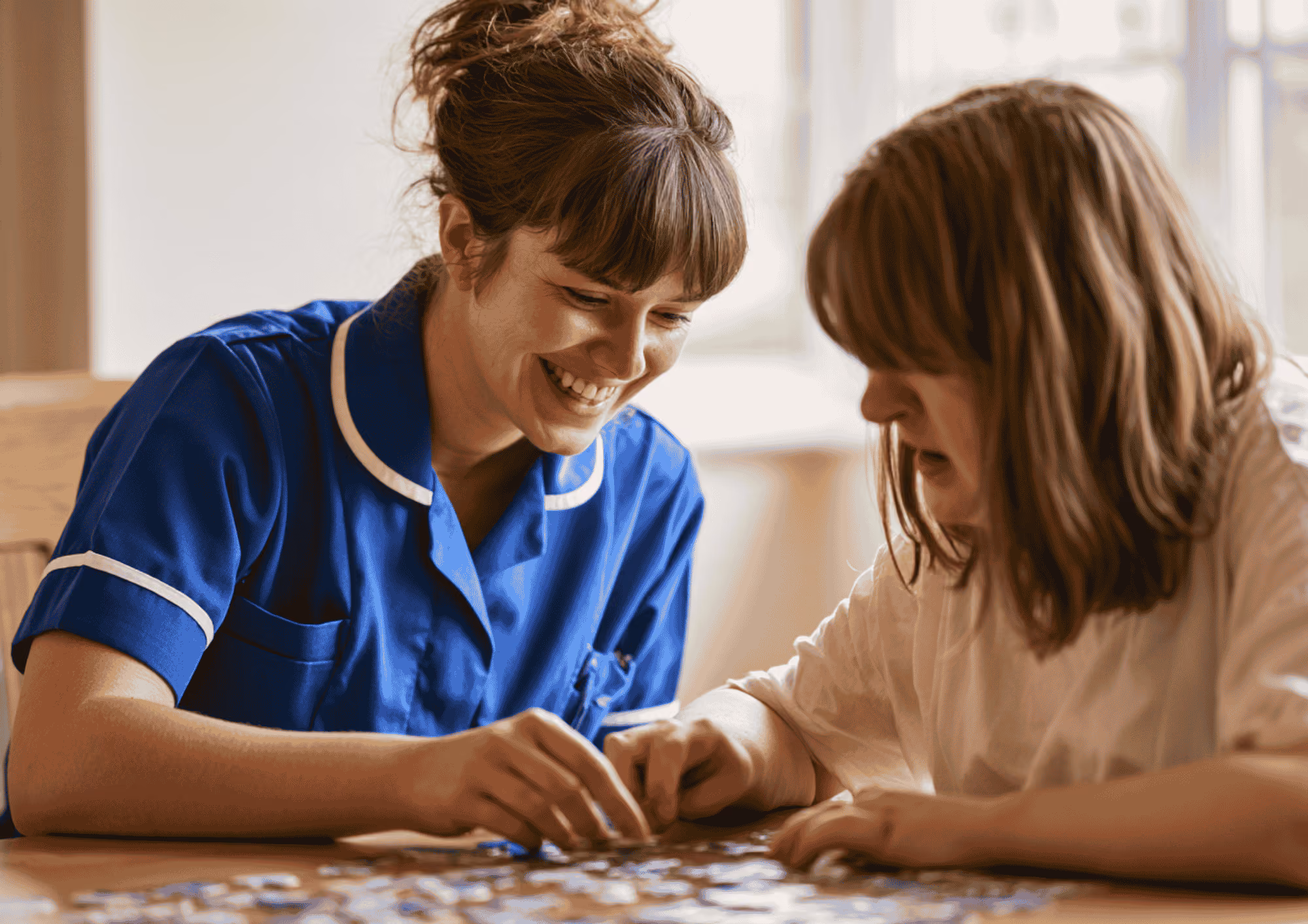 A smiling care worker in a blue uniform completing a jigsaw puzzle with a woman at home.
