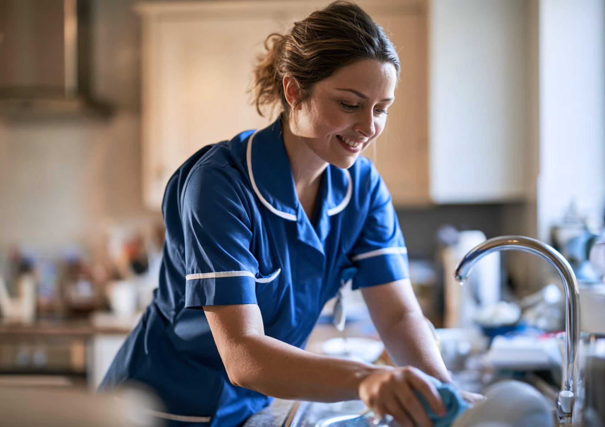 A care worker in a blue uniform washing dishes in a client’s kitchen.