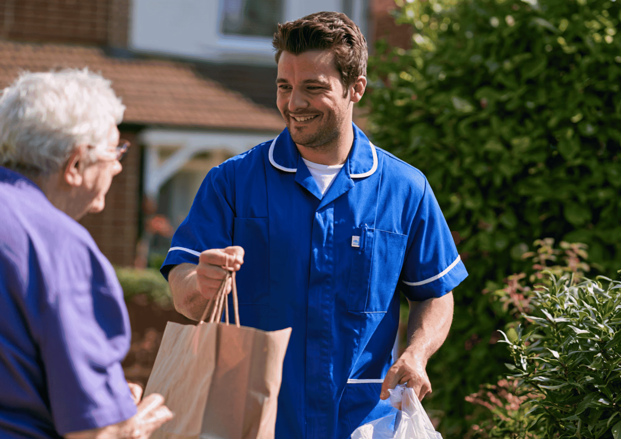 A smiling care worker in a blue uniform delivering shopping to an elderly woman at home.