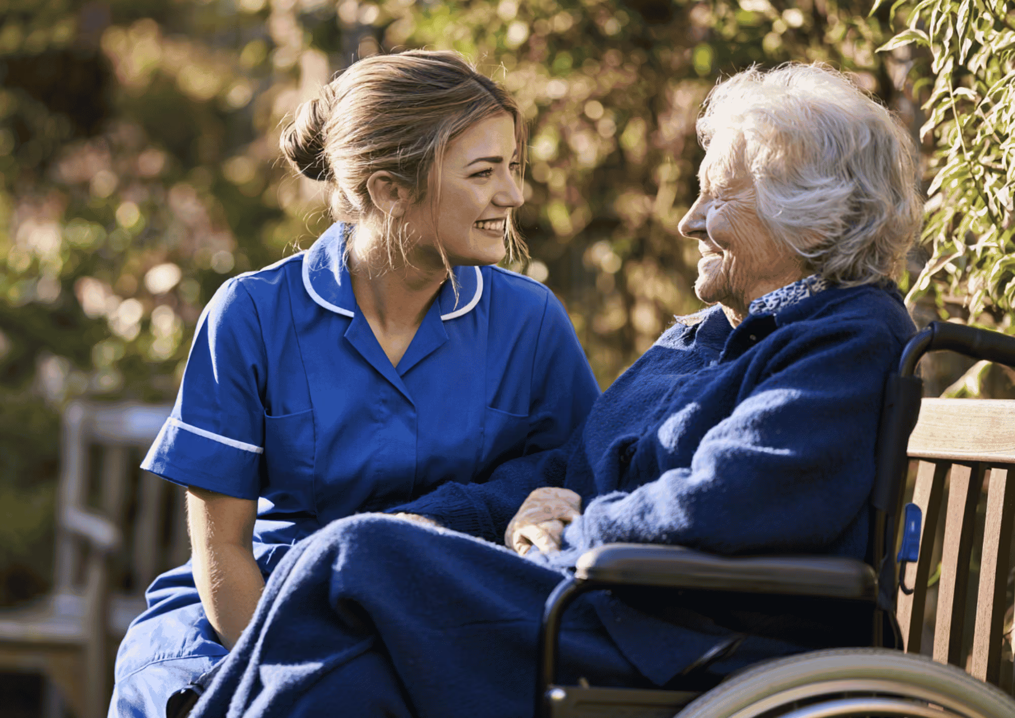 A smiling care worker in a blue uniform talking with an elderly woman using a wheelchair in a garden.