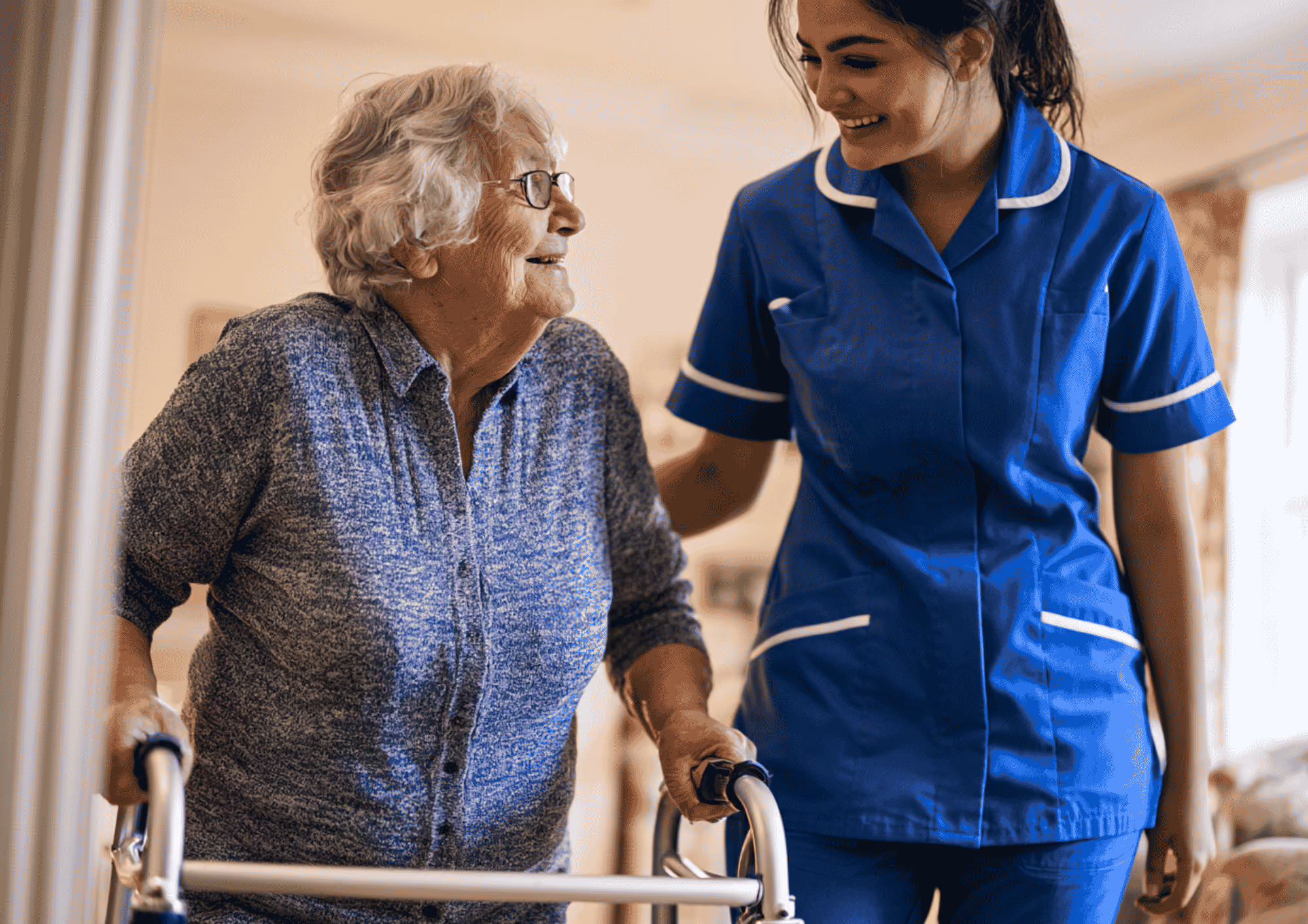 A smiling care worker supporting an elderly woman as she walks with a mobility frame at home.