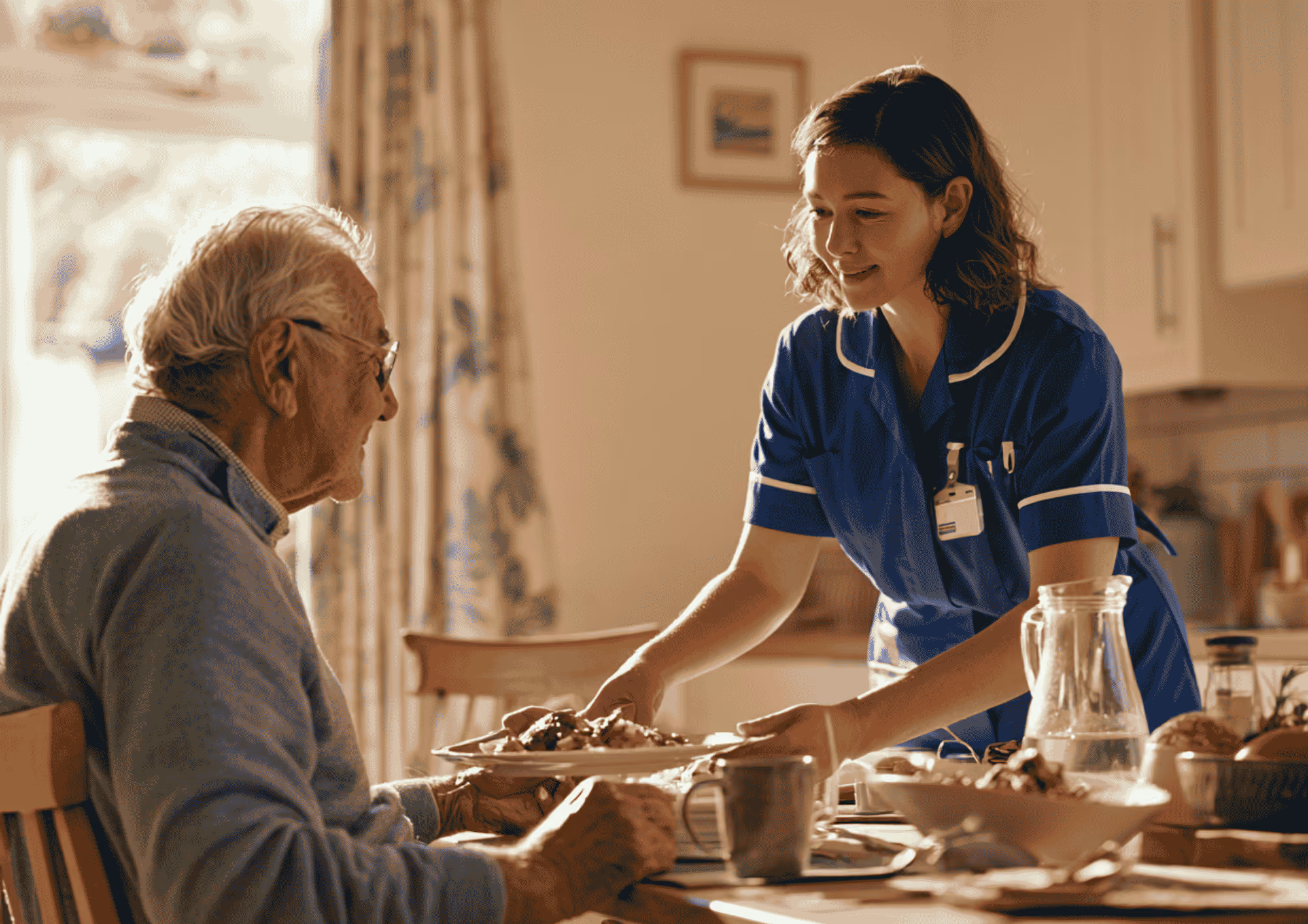 A care worker in a blue uniform serving a meal to an elderly man at his dining table.